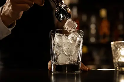 Providna čaša sa ledom dok barmen spušta kocku leda hvataljkom u ambijentu barskog osvetljenja